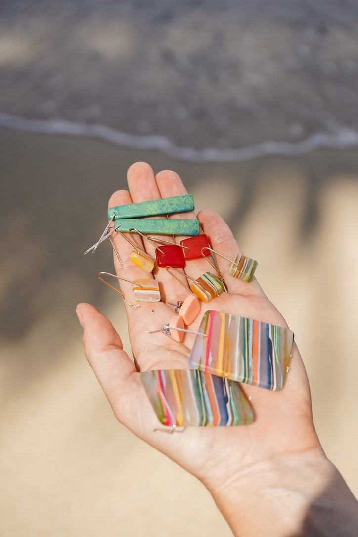 An open hand on the beach holding a pile of Surf Gems earrings in green, red, peach and striped rainbow resin, with the ocean behind