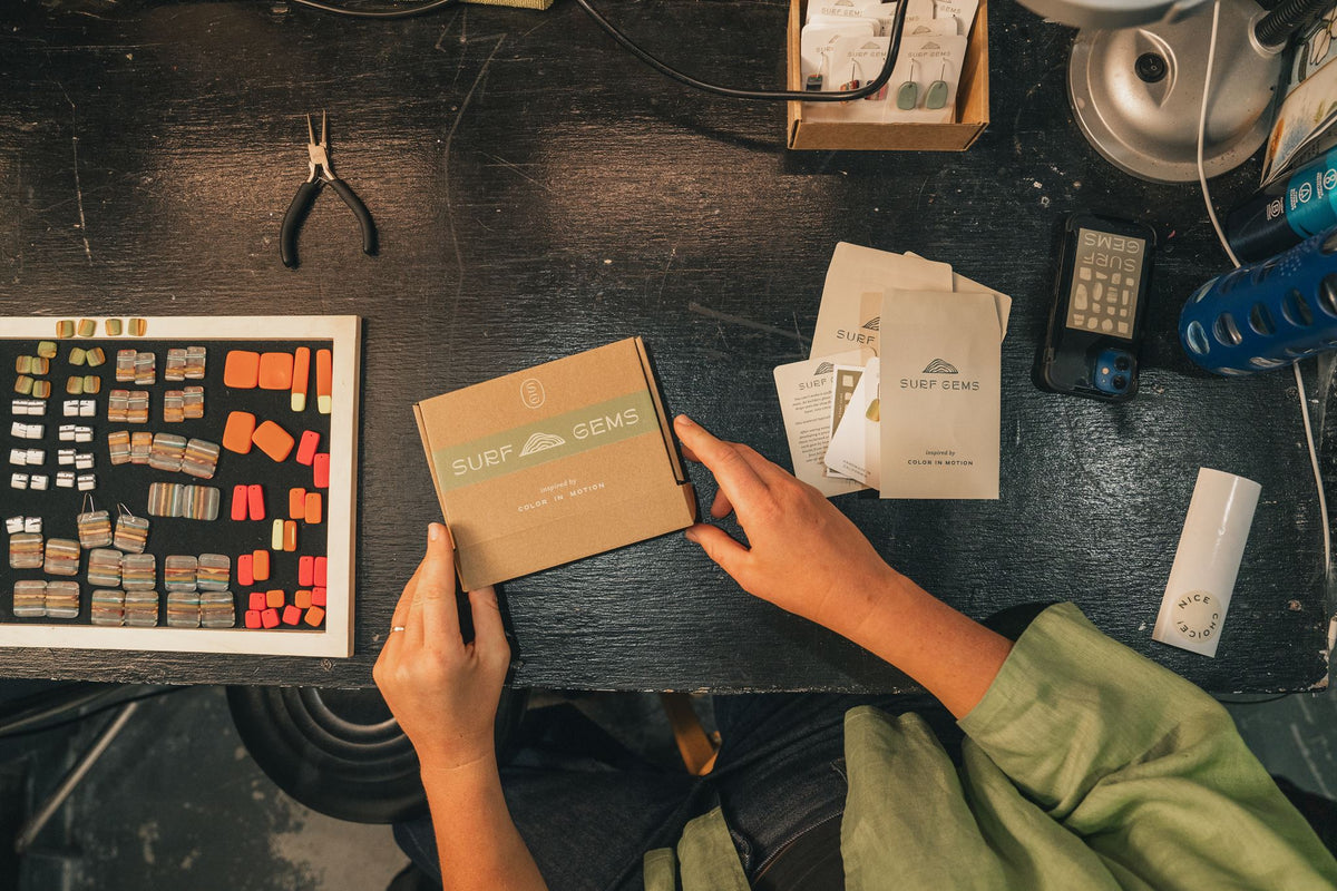 Hands holding a kraft Surf Gems gift box on a studio workbench, next to a tray of colorful resin earring pieces and Surf Gems earring cards